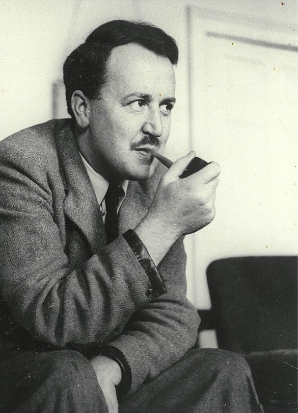 Black-and-white photograph of a man with a moustache seated indoors, wearing a textured suit jacket over a shirt and tie, holding a smoking pipe in one hand. A chair and a door are visible in the background.