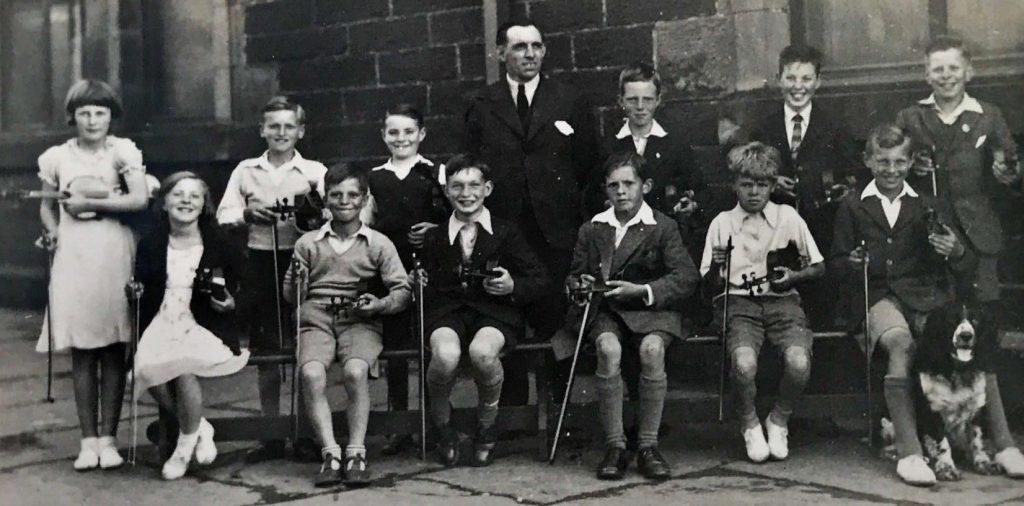 A black-and-white historical photograph showing a group of people posed outdoors in front of a stone building. The group consists of individuals seated and standing in two rows. Most of the seated individuals are holding violins and bows, suggesting a music class or orchestra group. A dog is sitting on the ground at the far right. The clothing appears formal and typical of the early 20th century, with suits, ties, and dresses.