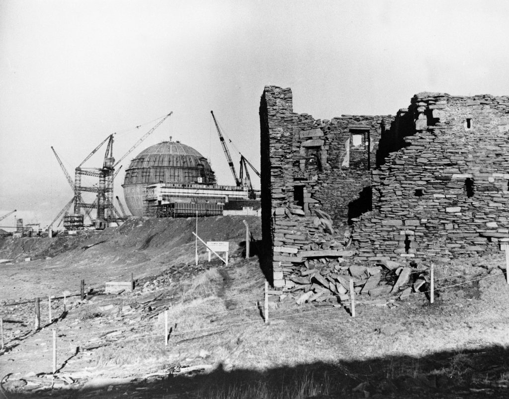 Black-and-white photo showing a partially ruined stone building in the foreground and the Dounreay nuclear reactor under construction in the background, with cranes surrounding the large spherical dome.