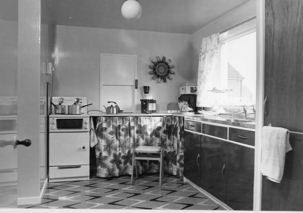 Black-and-white photo of a kitchen with stove, patterned curtain under counter, sink, window with floral curtains, and tiled floor.