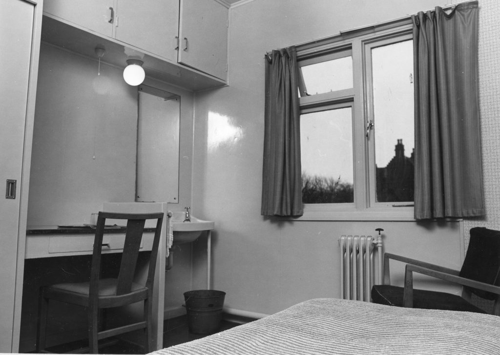 Black-and-white photo of a simple bedroom with bed, chair, desk and mirror, sink, radiator, and window with curtains.