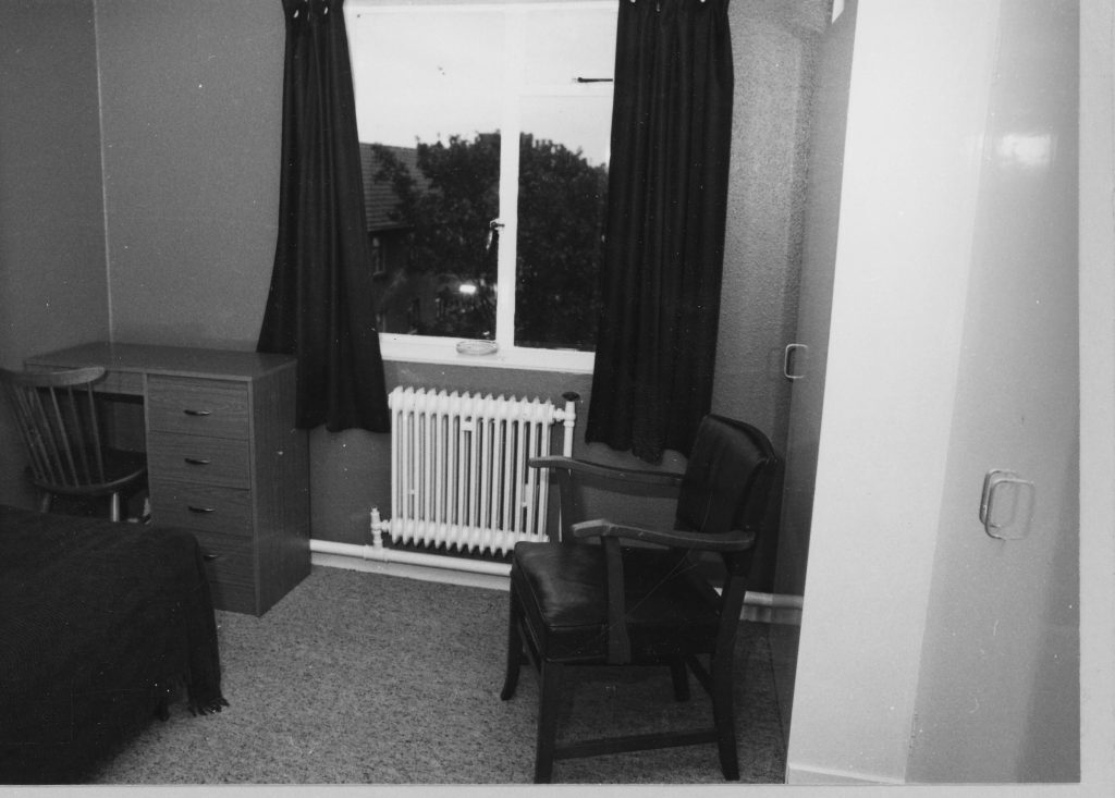 Black-and-white photo of a simple bedroom with bed, chair, chest of drawers, radiator, and window with dark curtains.