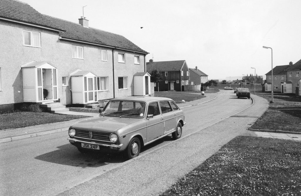 Black-and-white photo of a curved street with semi-detached houses and two parked cars, showing a quiet suburban housing estate.