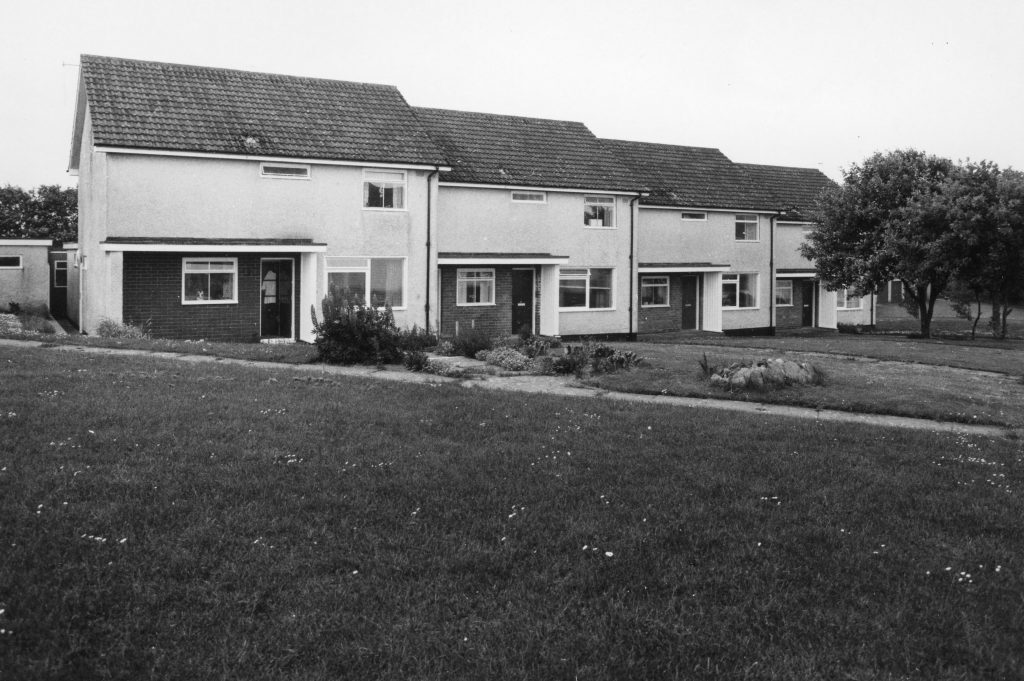 Black-and-white photo of terraced houses with pitched roofs and small front gardens, set behind a grassy area in a housing estate.