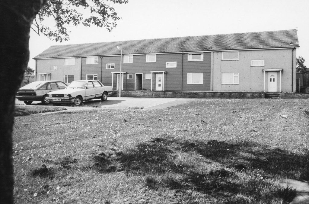 Black-and-white photo of a long row of terraced houses with pitched roofs, two parked cars, and grassy area in foreground.