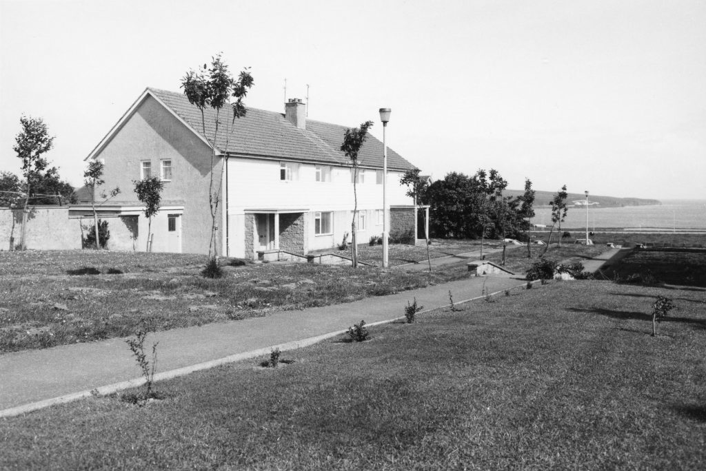 Black-and-white photo of a semi-detached house with pitched roof, grassy verge, street lamps, and distant view of coastline.