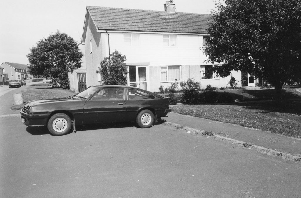 Black-and-white photo of a parked car in front of a semi-detached house with pitched roof, trees, and grassy verge in a housing estate.