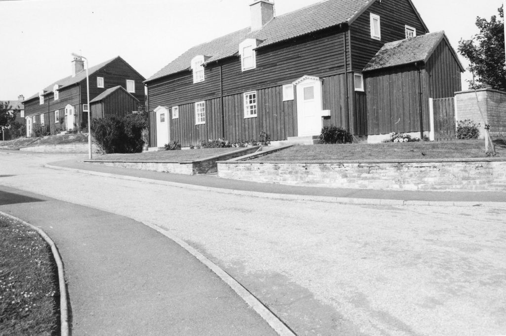 Black-and-white photo of a quiet street with large wooden-clad houses featuring pitched roofs and chimneys, set on raised plots.