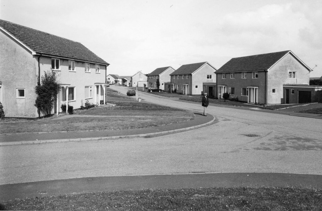 Black-and-white photo of a quiet street with semi-detached houses, grassy verges, and a person walking along the pavement.