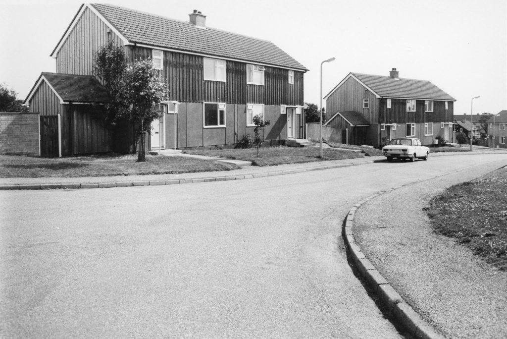Black-and-white photo of a quiet street with wooden-clad semi-detached houses, a parked car, and grassy verges under clear sky.