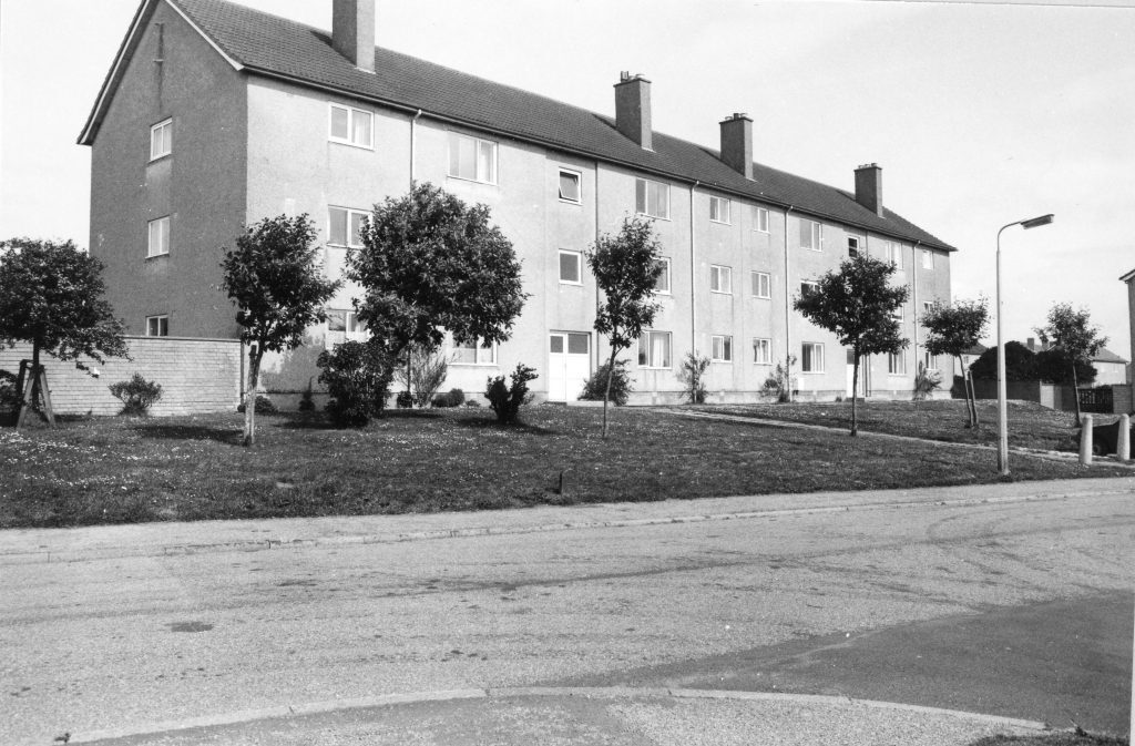 Black-and-white photo of a three-storey block of flats with pitched roof, chimneys, and small trees along a grassy verge by the road.