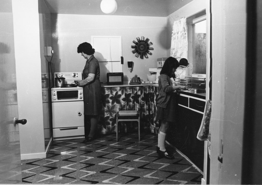 Black-and-white photo of a kitchen with patterned floor, stove, countertop appliances, and two people preparing food.