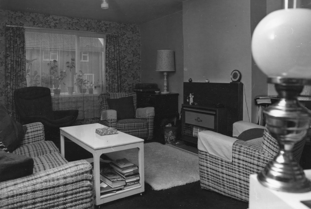 Black-and-white photo of a living room with patterned wallpaper, armchairs, sofa, coffee table, gas fireplace, and window.
