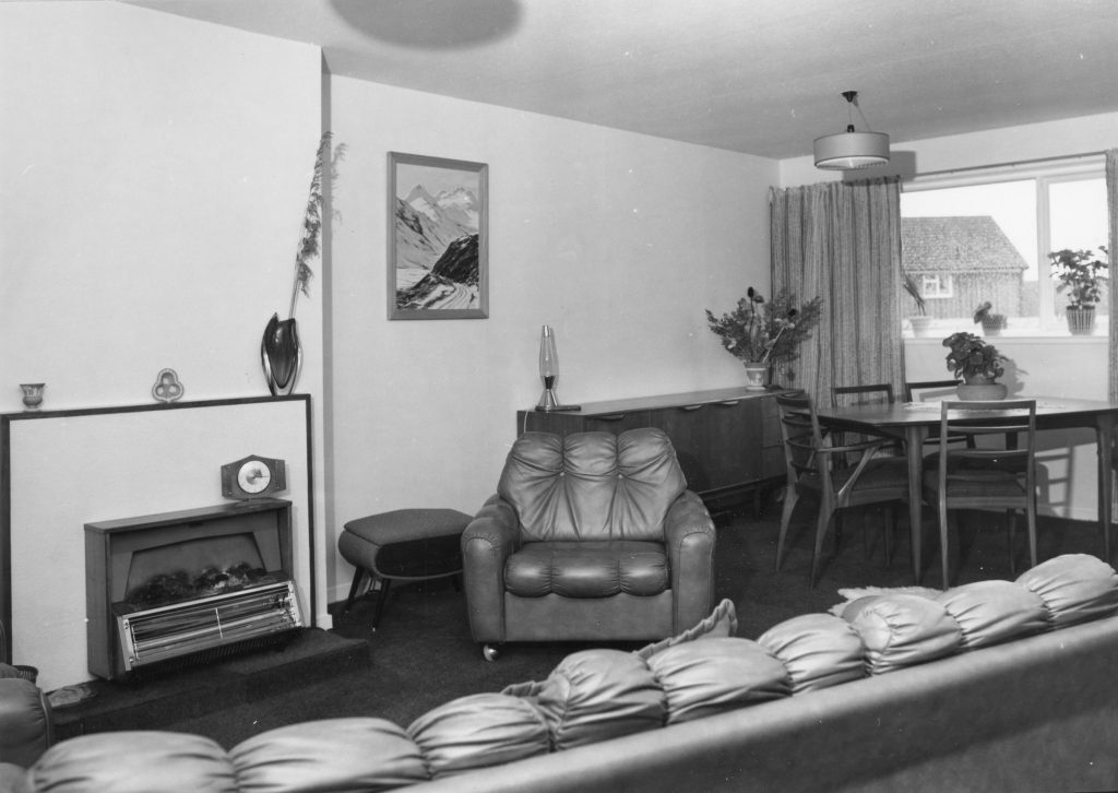 Black-and-white photo of a living room with armchair, sofa, fireplace, dining table, sideboard, and window with curtains.