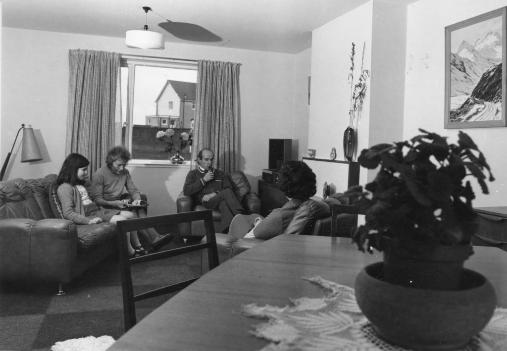 Black-and-white photo of a living room with armchairs, sofa, coffee table, wall art, and a window showing houses outside. Four people are sitting on the furniture.