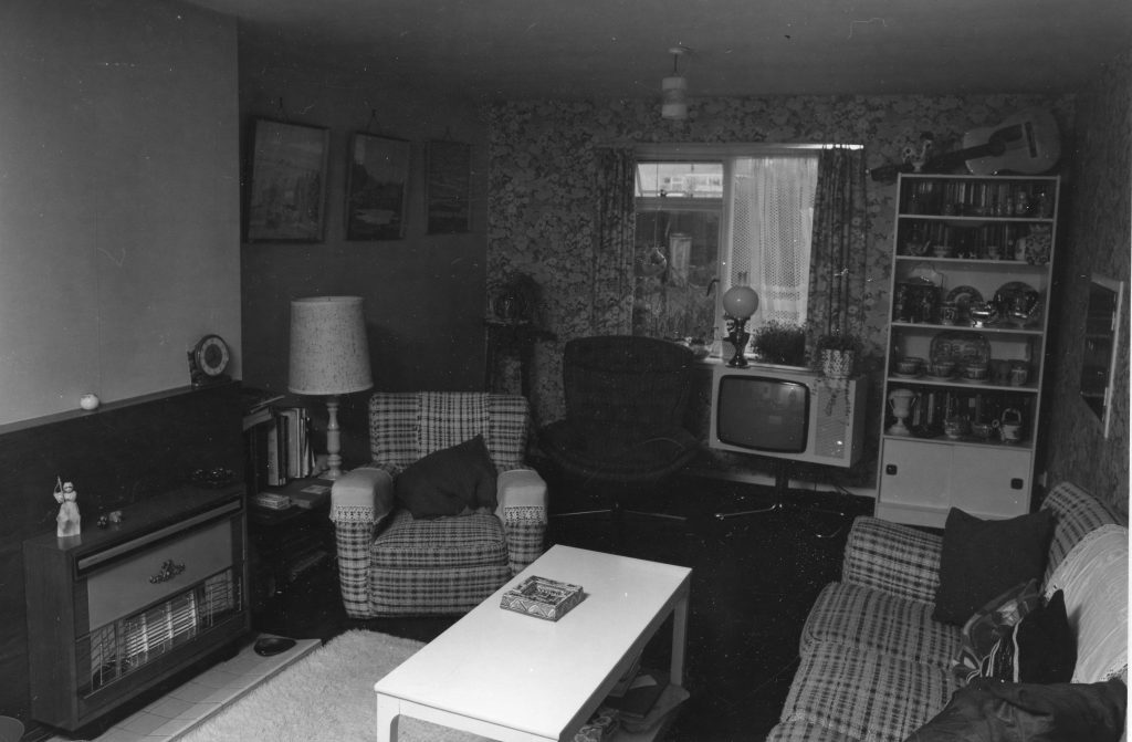 Black-and-white photo of a living room with patterned wallpaper, armchairs, sofa, coffee table, TV, bookshelf, and gas fireplace.