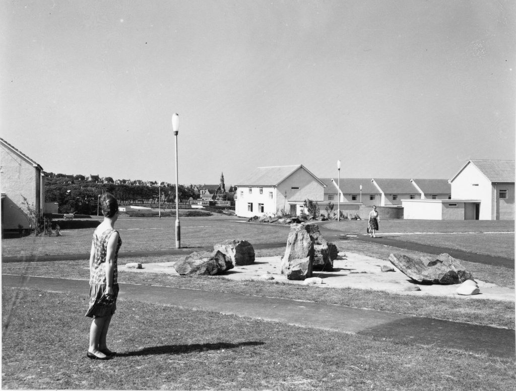 Black-and-white photo of a grassy housing estate with large rocks forming a feature, street lamps, and two women in 1950s/1960s style clothing walking near single-storey homes.