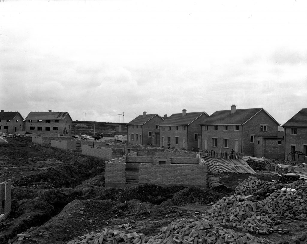 Row of partially built brick houses with piles of bricks and foundations on a muddy construction site under cloudy sky.