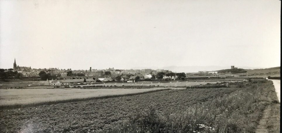 View of Thurso from a field, showing town buildings, church spire, and distant hills under a clear sky.