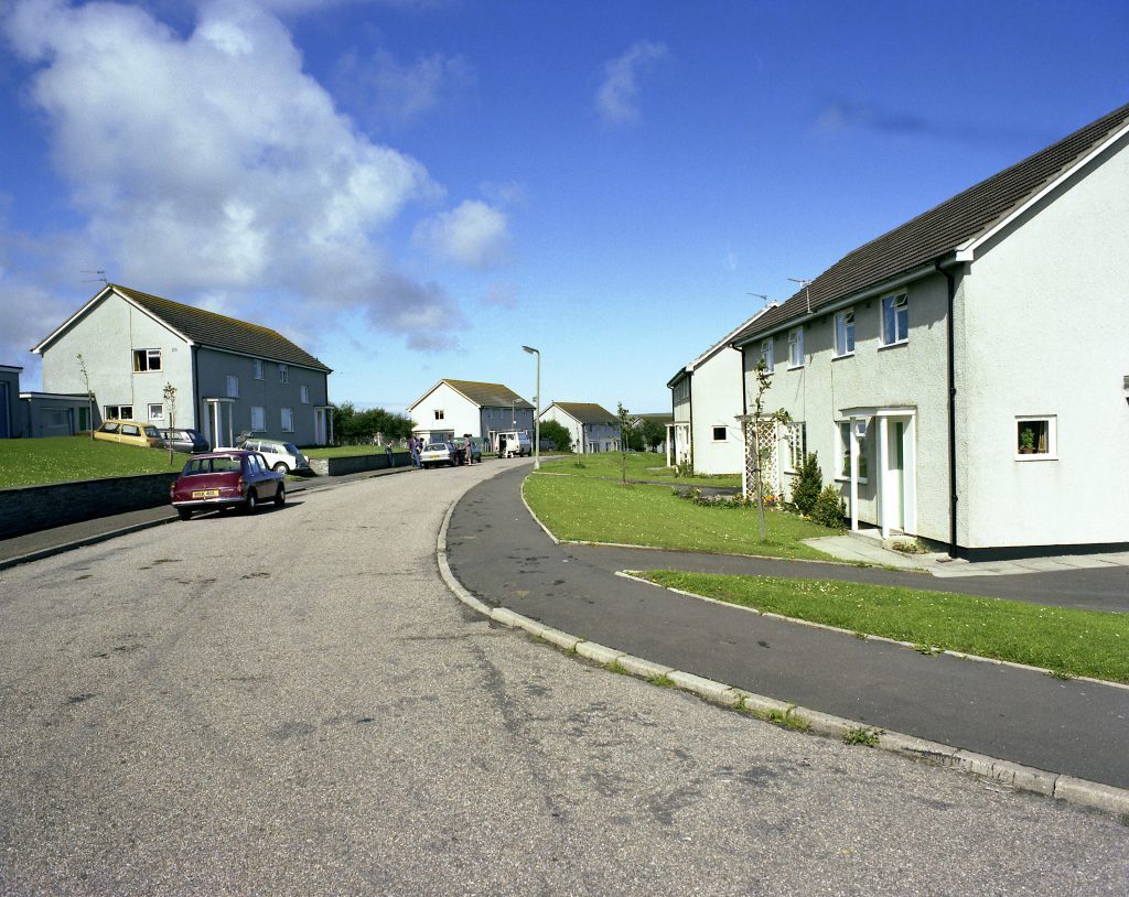 Curved street with semi-detached houses, green lawns, parked cars, and a bright blue sky with clouds.