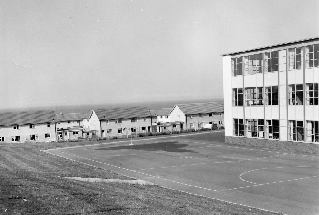 Black-and-white photo of a school building with large windows and a paved sports court in foreground. In the background are rows of houses.