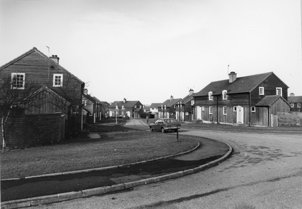 Black-and-white photo of a suburban street with wooden-clad houses, a parked car, and curved road in a housing estate.