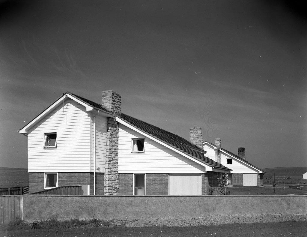Black-and-white photo of semi-detached houses with brick and timber walls, pitched roofs, and tall chimneys in a rural setting.