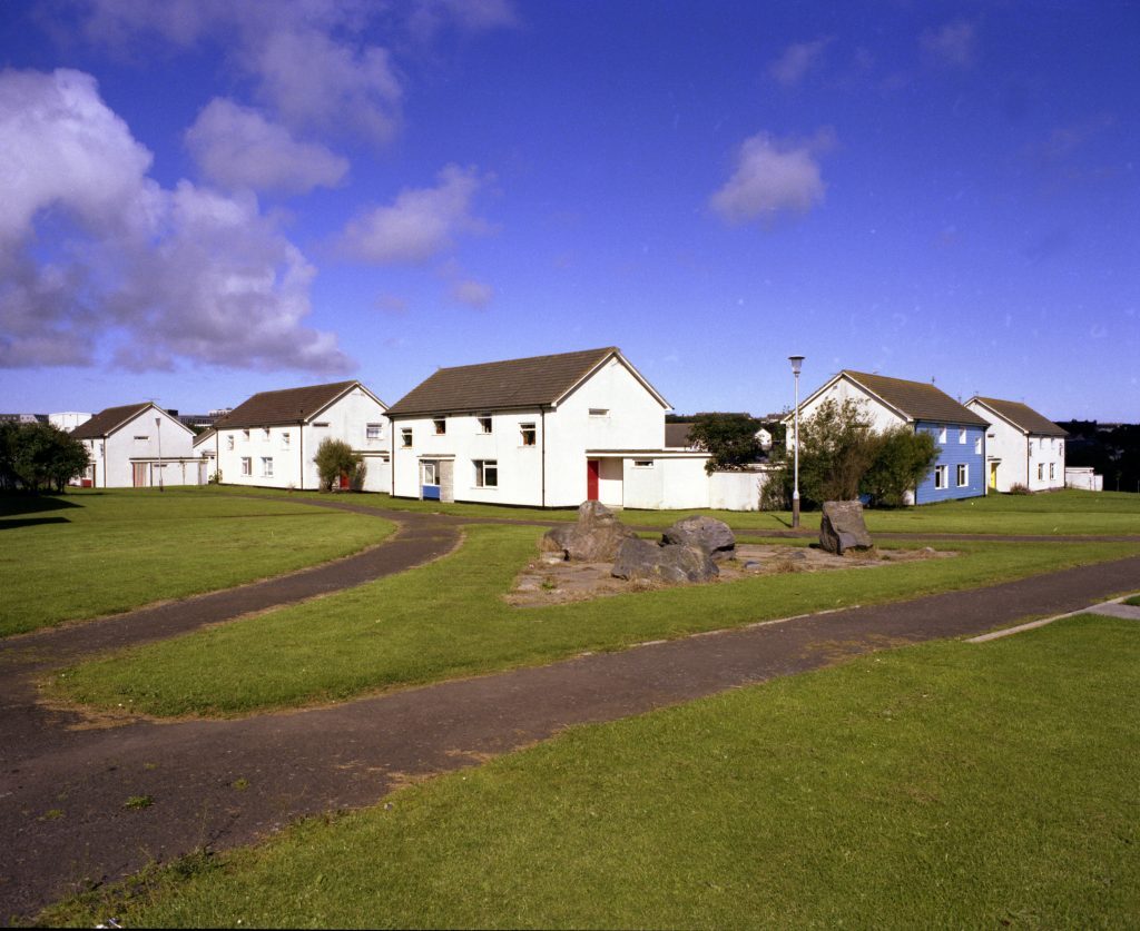 Row of modern white houses with pitched roofs and a blue house, set around grassy paths and rocks under a bright blue sky.