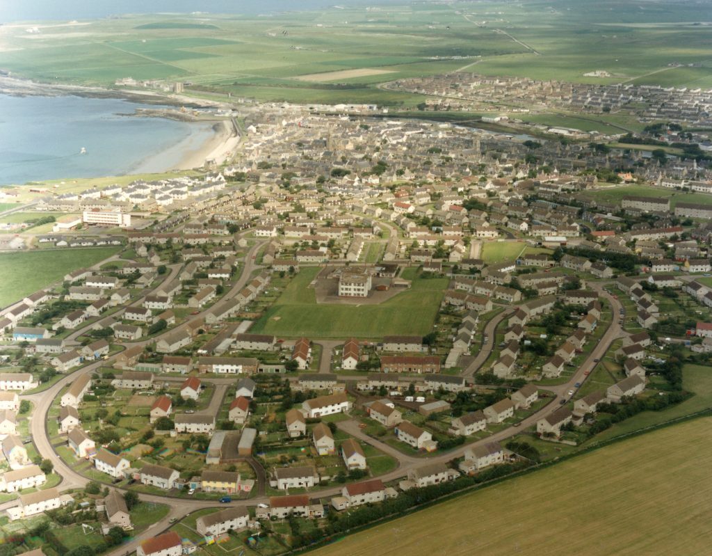 Aerial view of Thurso showing curved streets, houses, central building, coastline, and surrounding fields.