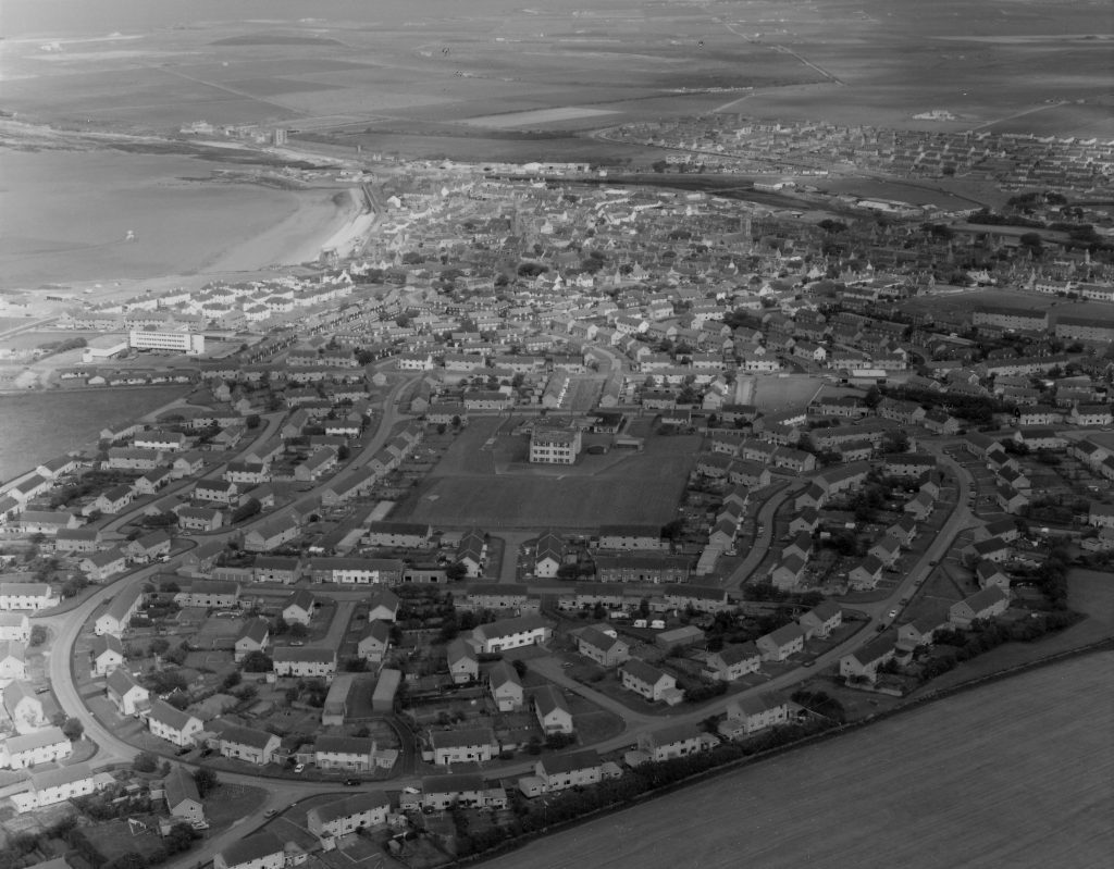 Aerial view of a coastal town with curved streets, houses, and open fields near the shoreline.