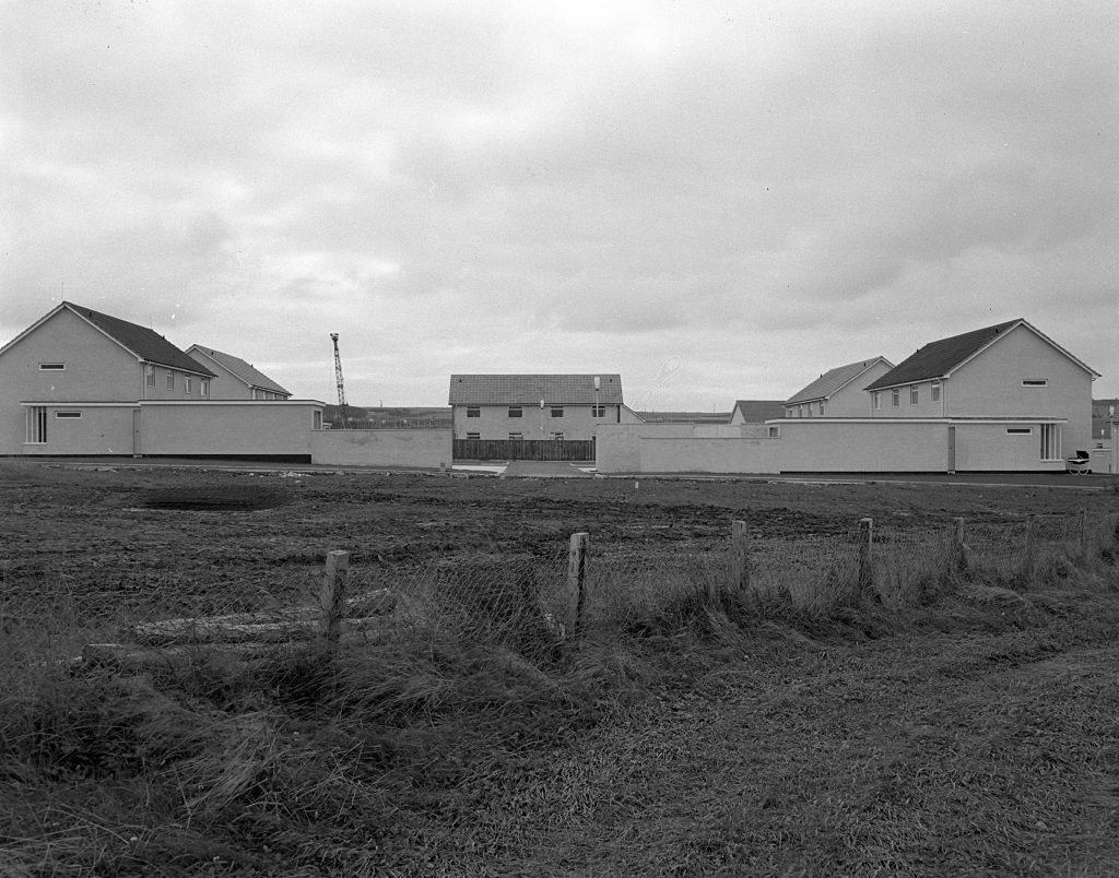 Partially built houses with white walls and pitched roofs on a cleared site, with fencing and open grassy foreground.