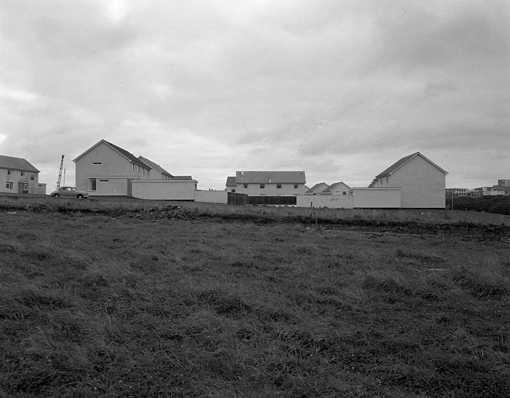 Partially built houses with white walls and pitched roofs on a cleared site, with grassy foreground and cloudy sky.