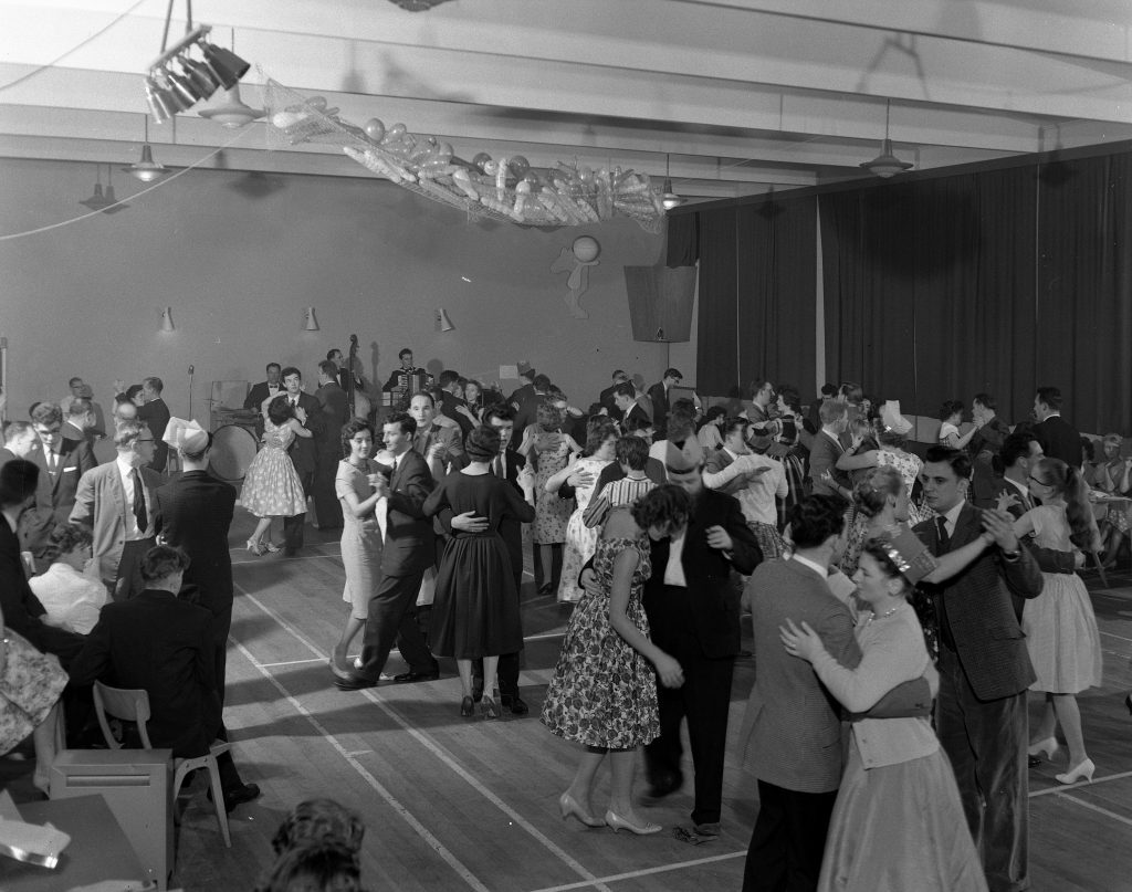 ack and white photograph of a crowded dance hall with numerous couples dancing in pairs on a polished wooden floor. The room is decorated with streamers and balloons hanging from the ceiling. A live band is visible in the background, playing instruments. Chairs and tables line the edges of the hall, where some people are seated.