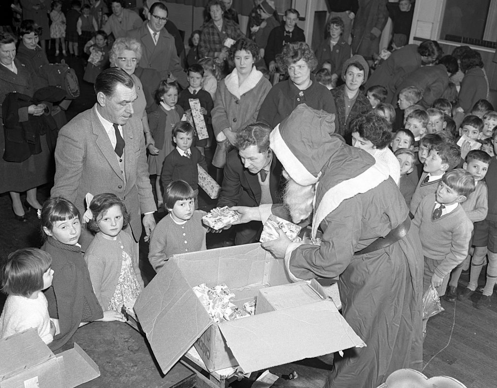 A festive gathering where a person dressed as Santa Claus distributes wrapped gifts from large cardboard boxes to a group of children. The children are standing in lines, some holding presents, while adults observe in the background. The setting appears to be an indoor hall with a wooden floor, and the scene suggests a mid-20th-century Christmas party.
