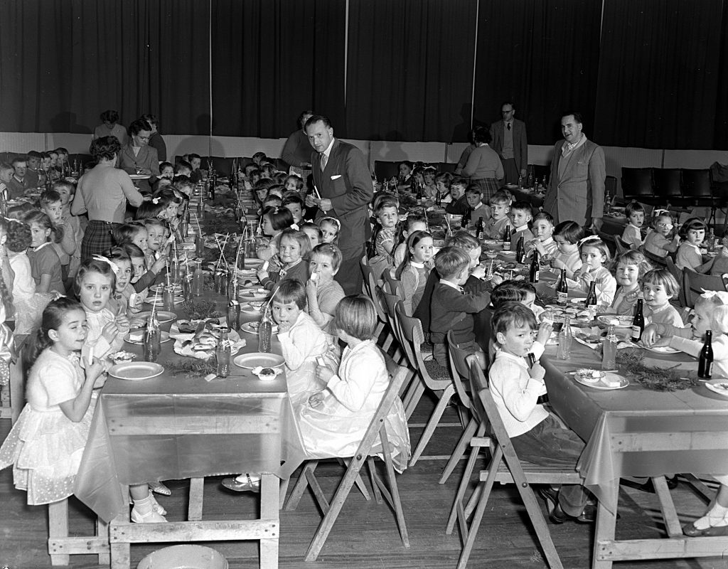 Large group of children seated at long tables in a hall, enjoying a festive meal. The tables are covered with tablecloths and set with plates, cutlery, bottles of drink, and decorations. Adults stand and move around the tables, assisting the children.