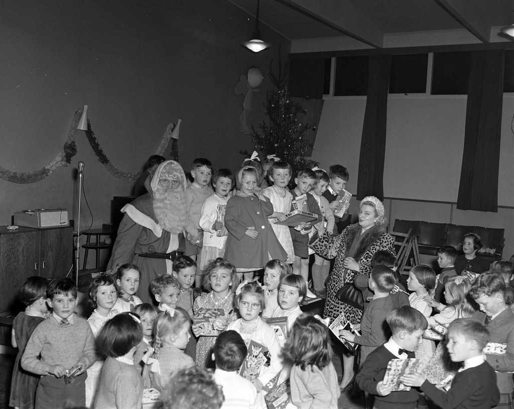 Black and white photograph of a children’s Christmas party in a hall. A person dressed as Santa Claus is seated near a decorated Christmas tree, surrounded by children holding wrapped presents. The room has festive decorations, including garlands on the walls and a microphone stand near a wooden cabinet. Rows of chairs are arranged along the side of the room.