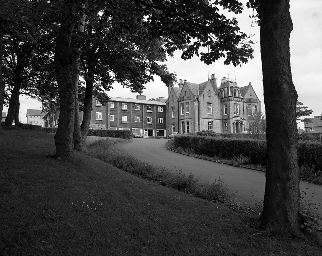 Black and white photograph of a large building complex featuring an older stone-built lodge with steep gabled roofs and chimneys, alongside a more modern rectangular extension with multiple windows. The buildings are set back from a curved driveway bordered by hedges, with a grassy slope and several tall trees in the foreground.