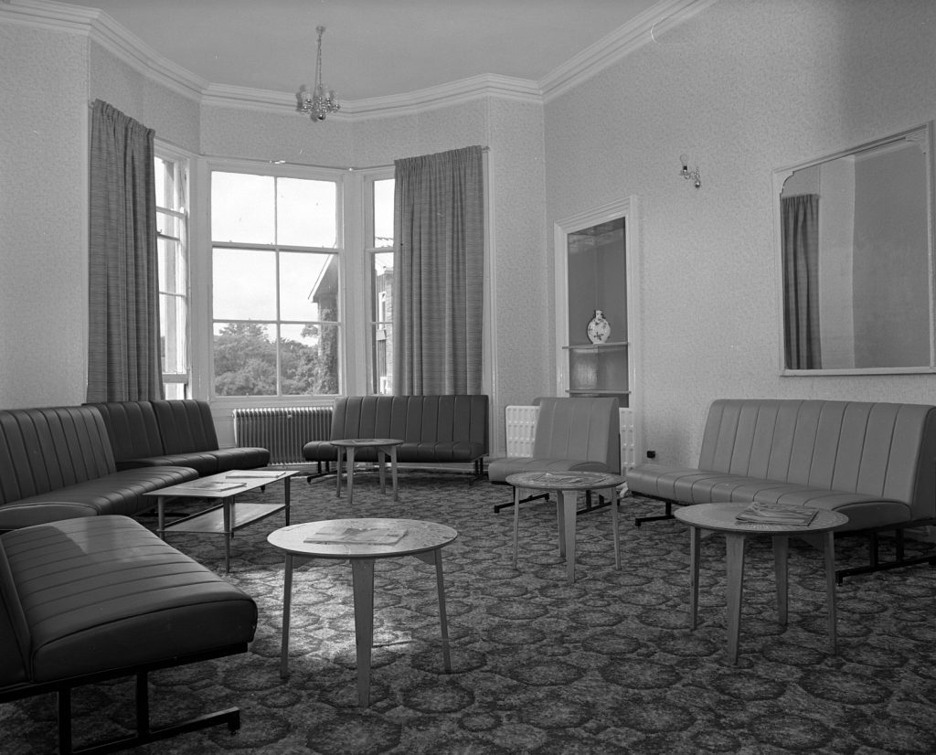 Black and white photograph of a lounge with patterned carpet and plain wallpaper. The room contains several armless upholstered sofas arranged around small circular tables. Large windows with curtains allow natural light into the space, and a decorative vase is displayed in a recessed wall niche. A rectangular mirror hangs on the wall, and two chandeliers are suspended from the ceiling.