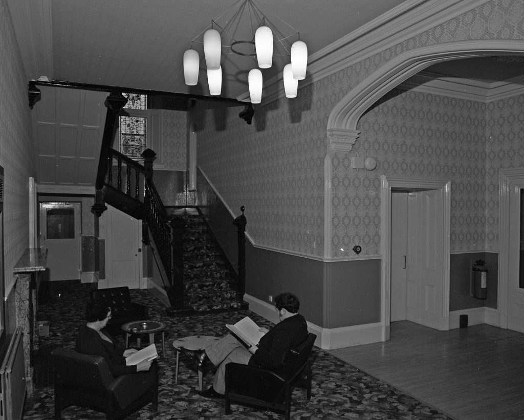 Black and white photograph of an interior hallway with patterned wallpaper and ornate architectural details. A staircase with a dark wooden banister leads upward on the left side, and a large arched doorway frames part of the space. Two people are seated in armchairs near a small round table, reading. A decorative light fixture with multiple pendant lamps hangs from the ceiling, and the floor is covered with a patterned carpet.