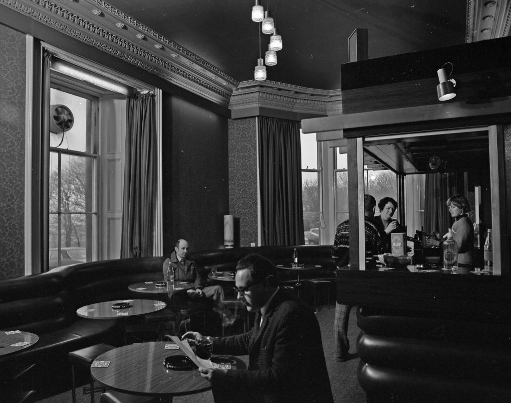 Black and white photograph of a lounge bar interior with ornate ceiling moldings and tall windows with curtains. The room has round tables and curved banquette seating along the walls. A wooden bar counter is on the right, where several people are standing and interacting with the bartender. Hanging pendant lights illuminate the space.