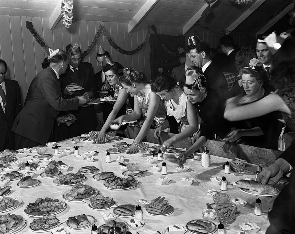 Black and white photograph of a festive buffet scene at a Christmas party. A long table is covered with a white cloth and set with numerous plates of food, including pastries, bread rolls, and other dishes, along with small condiment bottles and wrapped butter portions. Several people wearing party hats and formal attire are gathered around the table, reaching for food. The background shows wooden panel walls decorated with garlands and streamers under a sloped ceiling.