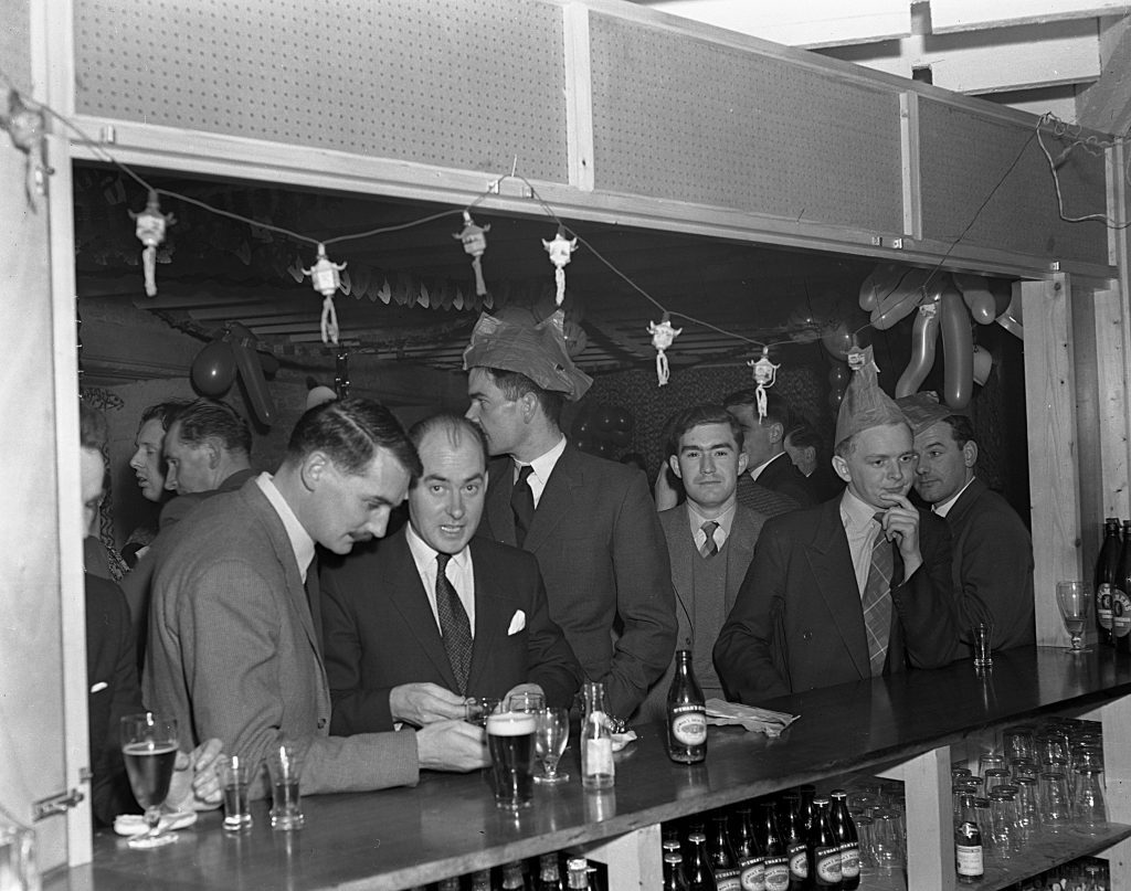 Black and white photograph of a bar scene during a festive event. Several people in suits and ties are gathered at the bar counter, which is lined with glasses and bottles of beer. Additional bottles are stored on shelves beneath the counter. The background is decorated with balloons, garlands, and hanging ornaments, and some individuals are wearing party hats.