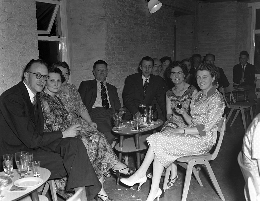 Black and white photograph of a group of people seated around small circular tables in a dimly lit room with stone walls. The tables hold several glasses and drinks, and the individuals are dressed in formal or semi-formal attire. The setting appears to be a social gathering or dance event, with simple wooden chairs and minimal decoration visible.