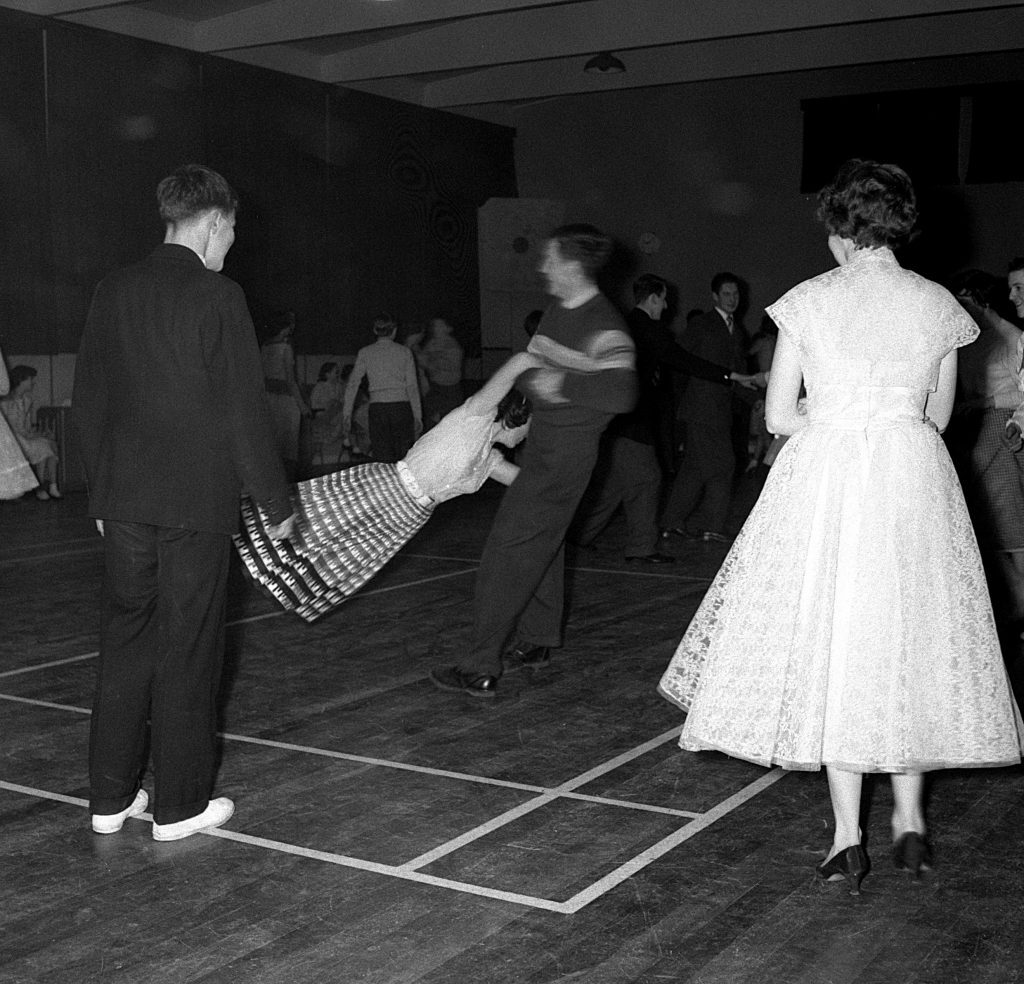 Black and white photograph of a lively dance scene in a hall with a wooden floor and plain walls. Several people are visible, with one person swinging another by the arms, causing the skirt to flare outward. Others stand nearby or dance in the background. The setting appears to be a social event, with individuals dressed in formal or semi-formal attire.