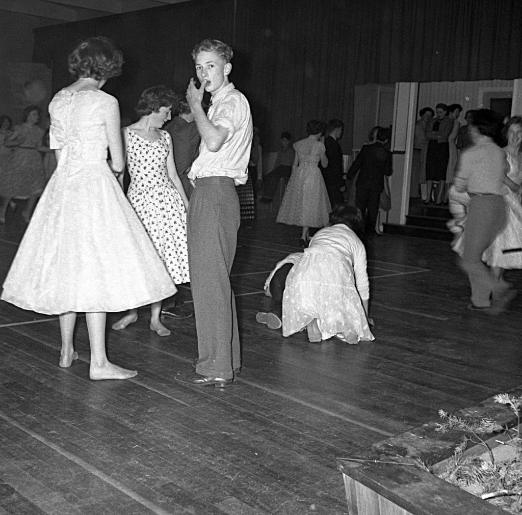 Black and white photograph of a dance hall with a polished wooden floor. Several people in formal attire are standing and talking, while others are dancing in the background. One person is kneeling on the floor near the center of the image, and a group of individuals can be seen gathered near a doorway at the back of the room. The setting includes plain walls with curtains and a raised platform area.