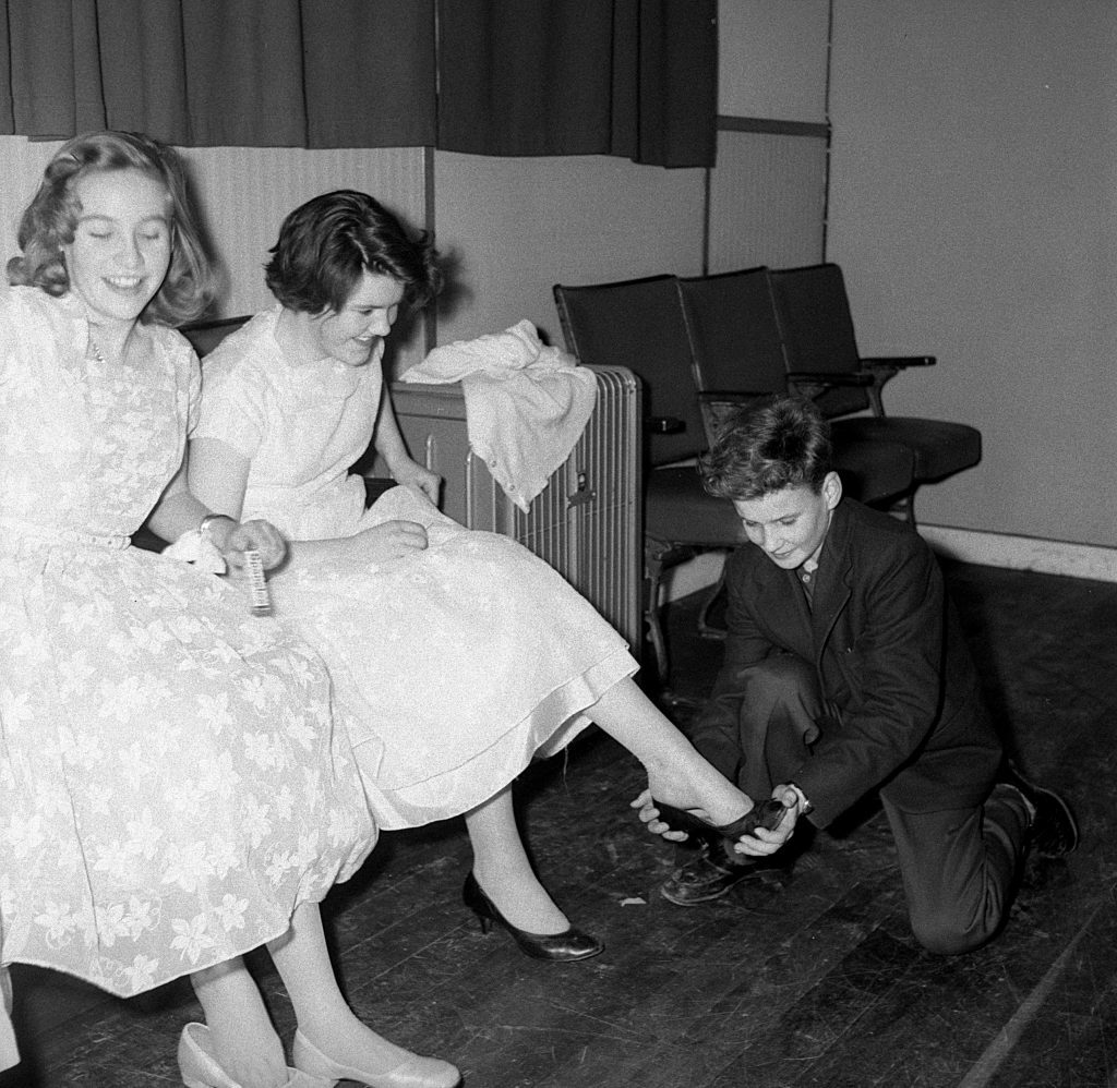 Black and white photograph showing two women seated in formal attire on chairs, with a young man kneeling on the floor adjusting a shoe on the foot of one of the women. A suitcase and folded garment are placed nearby, and a row of empty upholstered chairs lines the wall in the background.