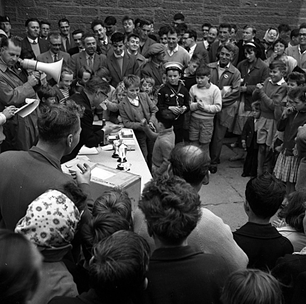 Prize-giving ceremony during a community event, with a large crowd gathered around a table placed against a brick wall. The table holds several trophies and awards, and an adult is handing a prize to a child at the front. People of various ages stand closely packed, watching the presentation, while some hold drinks or snacks. The setting appears outdoors on an overcast day.
