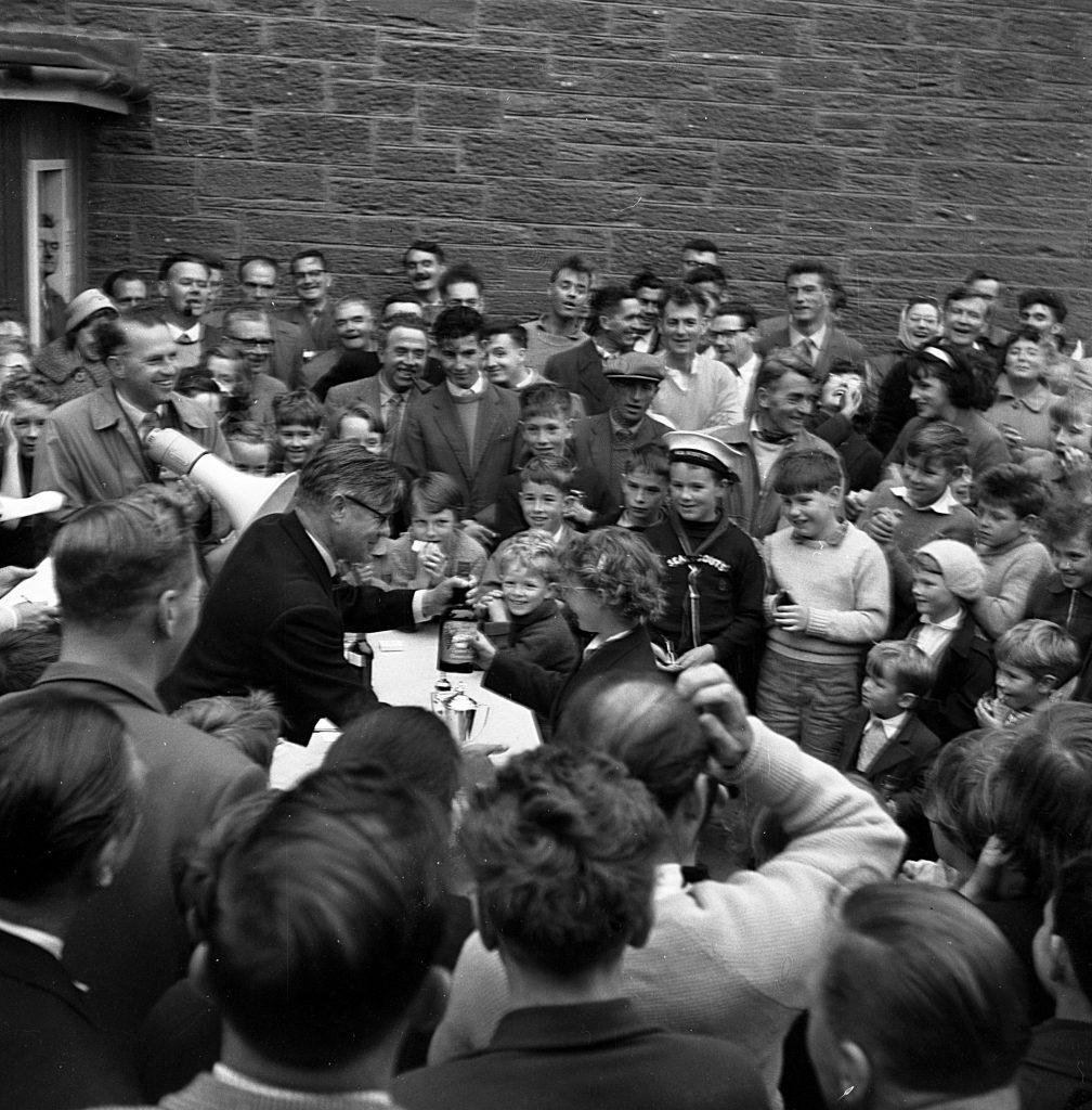 Prize-giving ceremony during a community event, with a large crowd gathered around a table placed against a brick wall. The table holds several trophies and awards, and an adult is handing a prize to a child at the front. People of various ages stand closely packed, watching the presentation, while some hold drinks or snacks. The setting appears outdoors on an overcast day.