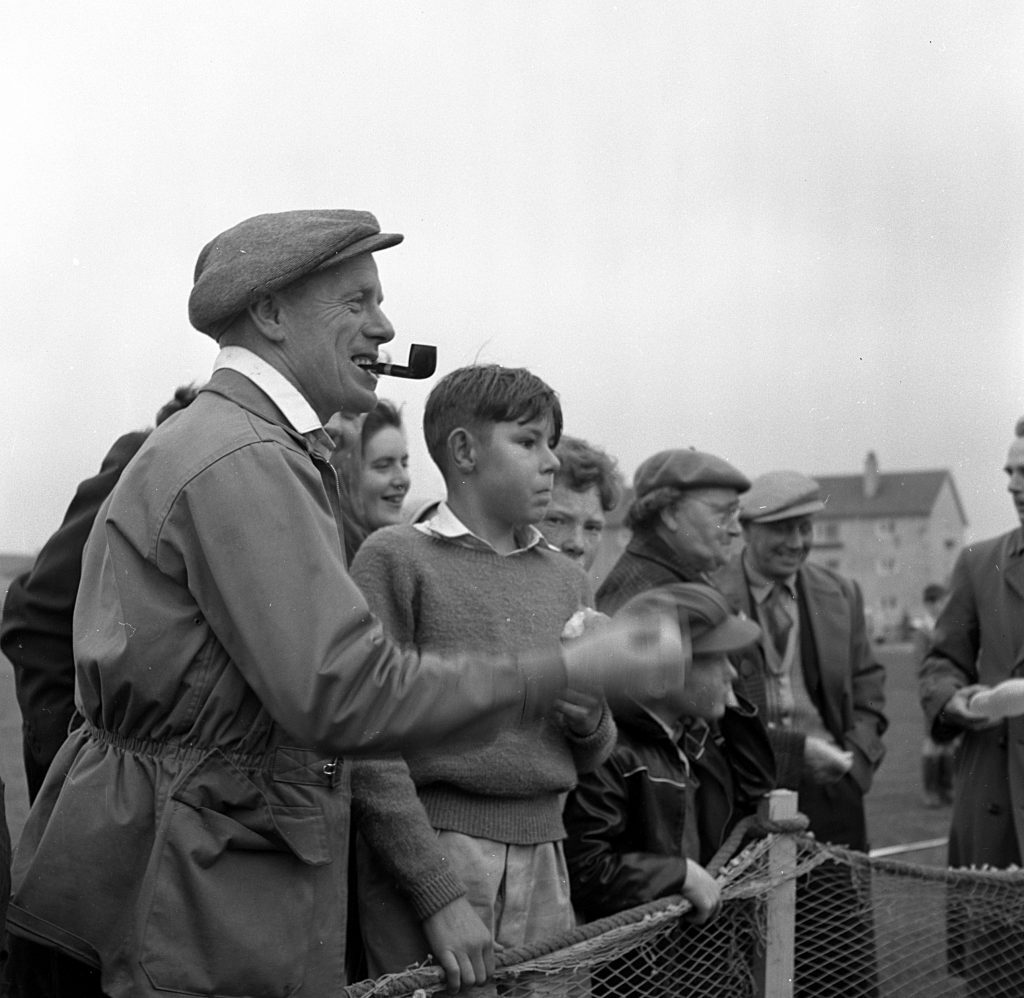 Group of spectators standing behind a rope barrier at an outdoor event on a grassy field. In the foreground, an older individual wearing a flat cap and a long coat is holding a pipe in his mouth and gesturing with one hand. Other people nearby are dressed in mid-20th-century clothing, including sweaters and jackets. In the background, houses and buildings are visible under an overcast sky.