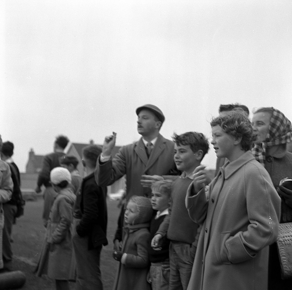 Group of spectators standing outdoors on a grassy field during a sports day event. Several people are dressed in mid-20th-century clothing, including coats, sweaters, and hats. One person in the center is possibly holding a small object and gesturing or throwing it, while others look toward the activity taking place off-frame. In the background, more individuals and buildings are visible under an overcast sky.